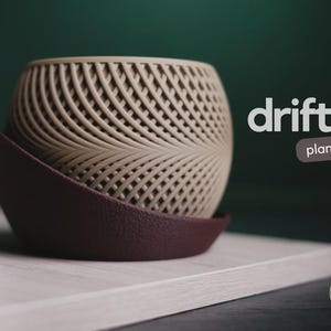 May include: A beige, spherical planter with a textured, repeating diagonal lattice pattern, resting inside a dark brown, curved base. The text "drift planter" appears in white and brown, and a small green logo reads "h3liø design".