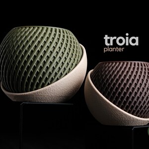 May include: Two Troia planter pots with a spherical shape and a textured, diagonal grid pattern. One planter is olive green and the other is dark brown, both set within a beige, rough-textured base. The text troia planter is visible.