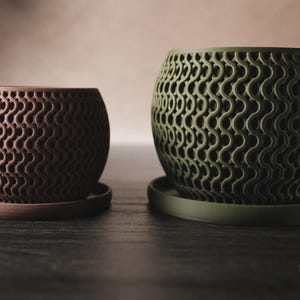 Puede incluir: Dos macetas redondas con un patr&oacute;n de celos&iacute;a ondulado, texturizado y repetitivo. Una maceta es de color marr&oacute;n oscuro y m&aacute;s peque&ntilde;a, mientras que la otra es de color verde oliva y m&aacute;s grande. Cada maceta se asienta sobre un plato circular a juego. Un logo dice h3liO.