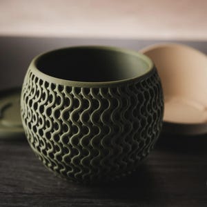 Puede incluir: Una maceta esf&eacute;rica de color verde oliva con un patr&oacute;n de celos&iacute;a ondulado repetitivo en su exterior. La maceta tiene una parte superior abierta y se acompa&ntilde;a de una base circular a juego en verde oliva y un cuenco de color beige claro.