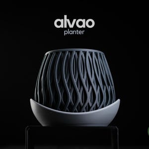 May include: A dark gray planter with a vertical wavy lattice pattern, resting in a light gray curved saucer. The text alvao planter is centered above the object, and a small green circular logo with h3liø design is in the bottom right corner.