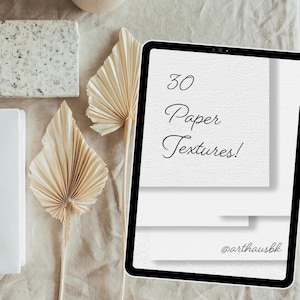 Könnte beinhalten: Ein digitales Tablet zeigt den Text "30 Paper Textures!" in einer Schreibschrift. Das Tablet ist von neutralen Dekorationselementen umgeben, darunter getrocknete Palmblätter und ein strukturiertes Element.