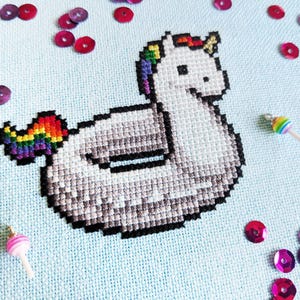 Può includere: Un disegno a punto croce di un unicorno bianco con criniera e coda arcobaleno, fluttuante su uno sfondo azzurro. L'immagine è decorata con paillettes rosse e viola e due ciondoli a forma di lecca-lecca.