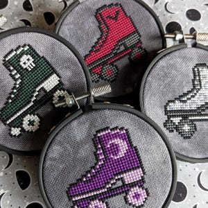 May include: Four embroidery hoops with cross-stitch designs of roller skates. The skates are in different colors: green, red, white, and purple.