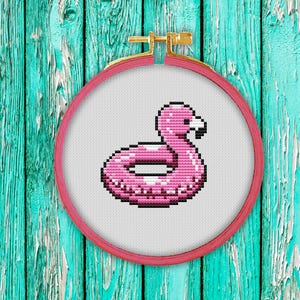 May include: Pink flamingo pool float cross-stitch pattern in a red embroidery hoop against a turquoise wooden background.