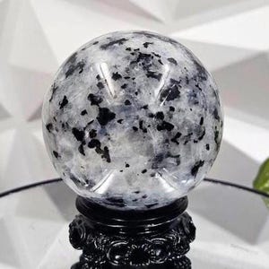 Puede incluir: Una esfera de cristal blanca y negra sobre un soporte negro. La esfera tiene una apariencia nublada y translúcida con motas negras en todo.