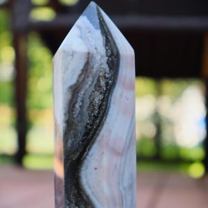 May include: A tall, polished crystal obelisk with a pointed top. The stone features a mix of colors, including white, gray, black, and pink, with swirling patterns and a textured appearance. The obelisk is a decorative item, likely for display or spiritual use.