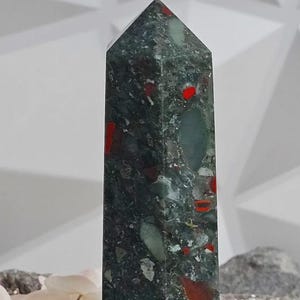 Könnte beinhalten: Ein hoher, rechteckiger Steinobelisk mit einer spitzen Spitze. Der Stein ist dunkelgrün mit roten und hellgrünen Einschlüssen. Der Obelisk steht aufrecht. Im Hintergrund sind weiße Blumen und eine helle Oberfläche zu sehen.