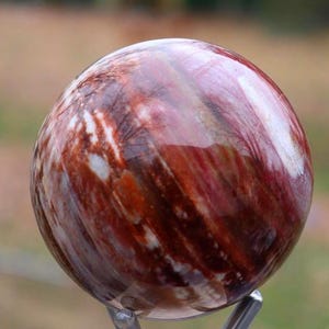 Pode incluir: Uma pedra esférica polida com um padrão de redemoinho de tons vermelhos, castanhos e brancos. A esfera parece estar montada em um suporte de metal, mostrando suas cores e texturas naturais. A superfície da pedra reflete a luz.