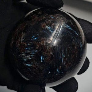Könnte beinhalten: Ein polierter, kugelförmiger Edelstein mit einer dunklen, fast schwarzen Grundfarbe. Der Stein weist komplizierte Muster aus braunen und blaugrünen Streifen und Einschlüssen auf, die ein einzigartiges, strukturiertes Aussehen erzeugen. Die Kugel wird vor einem dunklen Hintergrund gehalten.