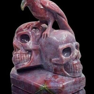 May include: A carved sculpture of two skulls and a raven, in a deep red colour. The raven is perched on the skulls, with detailed feather and beak carvings. The base is rectangular, and the piece has a polished finish.