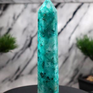 May include: A tall, polished crystal obelisk with a pointed top. The obelisk is a vibrant teal colour with darker green and black inclusions. The crystal is standing upright on a black surface.