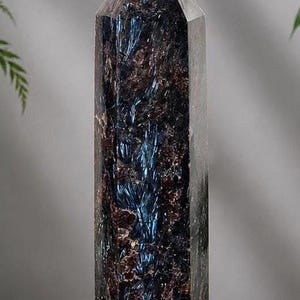Puede incluir: Una torre de cristal alta de seis lados con una punta afilada. La piedra presenta una superficie oscura y texturizada con tonos marrones y negros profundos, acentuada por llamativas vetas iridiscentes de azul metálico que corren.