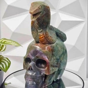 May include: A green and purple stone skull sculpture with a green and orange stone snake on top. The sculpture is carved in detail and has a realistic look.