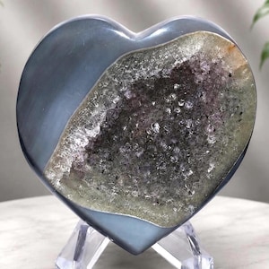 Puede incluir: Una escultura de piedra pulida en forma de corazón con una superficie exterior lisa de color gris oscuro y una cavidad de geoda central llena de pequeños cristales de amatista de color púrpura brillante sobre un soporte de plástico.