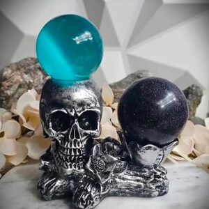 May include: A silver skull-shaped sculpture holds a teal-colored sphere on top and a dark blue sphere in a decorative holder. The sculpture has intricate details and is set against a neutral background with floral accents.