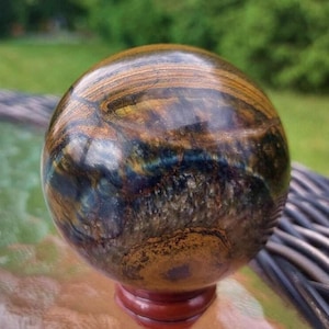 May include: A polished sphere with a swirling pattern of brown, gold, and blue hues. The orb is mounted on a small, reddish-brown wooden stand, showcasing the natural colors and textures of the stone.