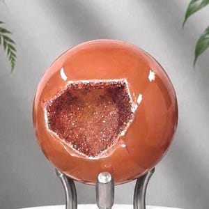 May include: A polished, spherical orange stone featuring a central geode cavity filled with small, sparkling crystals. The smooth, rounded surface of the sphere contrasts with the textured, crystalline interior. It rests on a metal stand.