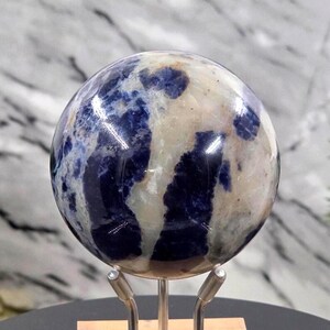 May include: A polished sphere of sodalite, showcasing a mix of deep blue and white hues. The gemstone is mounted on a silver-toned metal stand with a wooden base, creating a decorative display piece.
