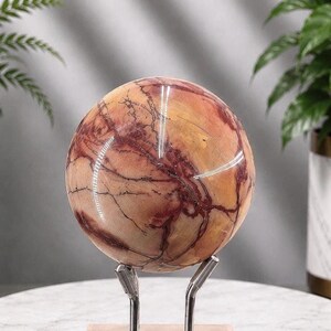 May include: A polished spherical stone featuring a warm colour palette of tan, cream, and deep red veins. The sphere is supported by a silver-toned metal stand with two prongs that hold the object securely in place, showcasing its smooth, glossy surface.
