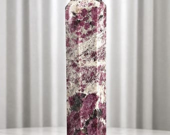 Könnte beinhalten: Ein hoher, sechseckiger Kristallturm mit polierter Oberfläche und einem gesprenkelten Muster aus tief magentafarbenen, weißen und dunkelgrauen Mineraleinschlüssen. Der Stein hat eine facettierte Spitze und eine flache Basis mit natürlicher Textur.