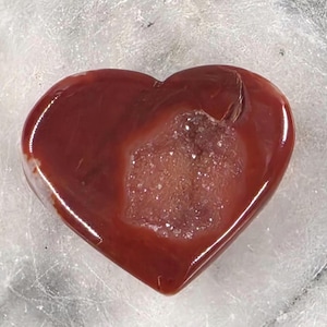 Puede incluir: Una gema pulida en forma de coraz&oacute;n, en tonos rojos profundos con un centro cristalino m&aacute;s claro. La piedra tiene una superficie lisa y brillante, probablemente un objeto decorativo o una joya.