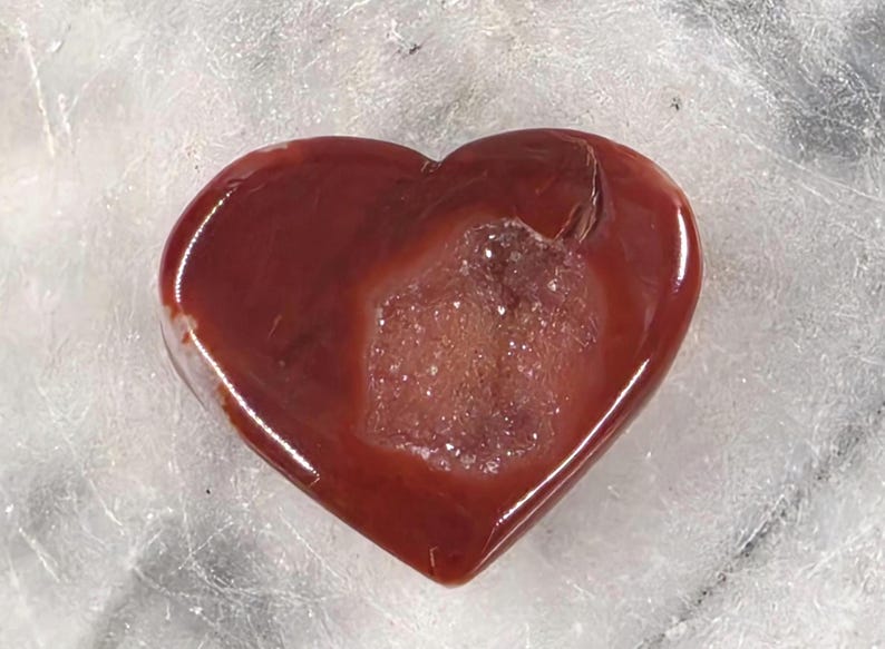 Puede incluir: Una gema pulida en forma de coraz&oacute;n, en tonos rojos profundos con un centro cristalino m&aacute;s claro. La piedra tiene una superficie lisa y brillante, probablemente un objeto decorativo o una joya.