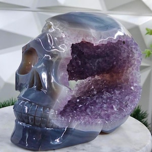 May include: A carved skull sculpture made of polished agate stone. The skull is a gradient of gray and blue, with a cavity filled with sparkling purple amethyst crystals. The sculpture is displayed on a white marble surface.