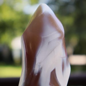 May include: A polished stone sculpture with a smooth, flame-like shape. The surface features a distinct banded pattern with alternating layers of translucent white and deep reddish-brown, creating a natural, flowing contrast across the entire object.