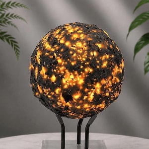 May include: A spherical lamp with a textured, dark charcoal-colored exterior featuring irregular openings that emit a warm, bright yellow light. The orb is supported by a black metal stand with three prongs resting on a clear square base.