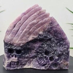 以下が含まれることがあります： 紫とラベンダー色の彫刻された石の彫刻で、様式化されたスカルのデザインが特徴です。彫刻はテクスチャ加工された表面とユニークな芸術的な外観を持っています。スカルの特徴が明確に定義されており、作品の複雑なディテールを加えています。