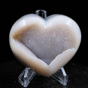May include: A heart-shaped geode with a polished, pearly white exterior and a sparkling, crystal-filled interior. The geode is displayed on a clear acrylic stand, showcasing its natural beauty. The interior features shades of gray and brown.