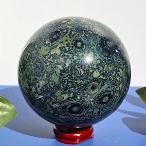 Könnte beinhalten: Poliertes, kugelförmiges Dekorationsobjekt mit einem dunkelgrünen und schwarzen, gesprenkelten Muster. Die Kugel steht auf einem kleinen, runden, roten Holzsockel. Die Oberfläche hat ein glänzendes Finish und ein einzigartiges, organisches Aussehen.