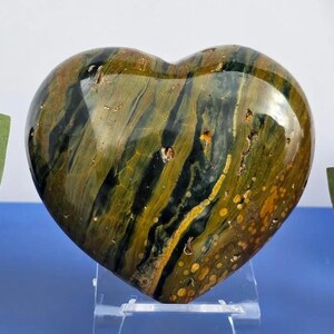 Könnte beinhalten: Ein polierter herzförmiger Stein mit einer Mischung aus grünen, gelben und braunen Farbtönen. Der Stein hat eine glatte, glänzende Oberfläche und wird auf einem transparenten Ständer präsentiert. Die Herzform ist prominent.