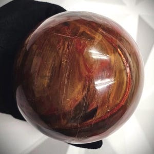 Könnte beinhalten: Ein polierter, kugelförmiger Edelstein mit einer reichen, erdigen Farbpalette. Die Kugel weist wirbelnde Muster aus tiefem Braun, Rotbraun und Goldtönen auf. Die Oberfläche hat eine glänzende Oberfläche, die das Licht reflektiert und die natürliche Maserung des Steins zur Geltung bringt.