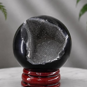 May include: A polished black stone sphere featuring a large, textured, crystalline geode cavity on its front surface. The sphere is supported by a circular, glossy, dark red wooden stand with a tiered, rounded design and smooth finish.
