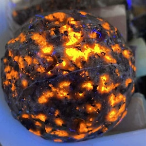 May include: A close-up of a glowing, round rock with a dark gray exterior and vibrant orange and yellow interior. The rock has a rough, textured surface with small blue highlights. Other rocks are visible in the background.