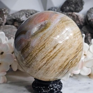 May include: A large, polished sphere with a swirling pattern of tan, brown, and pink hues. The orb is mounted on a black, ornate stand. The sphere's surface has a glossy finish, reflecting light.