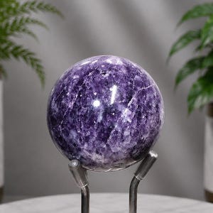 May include: A polished amethyst sphere featuring deep purple hues with white crystalline inclusions and natural fractures throughout the stone. The round crystal is displayed resting on a silver-toned metal stand with two curved support arms.