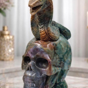 May include: A carved stone sculpture featuring a cobra snake coiled atop a human skull. The piece displays a marbled texture with variegated hues of deep green, brown, blue, and purple throughout the polished surface of the stone material.