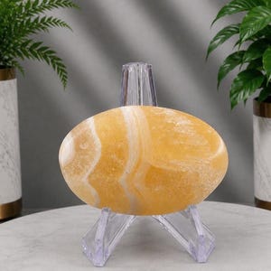 May include: A smooth, oval-shaped yellow stone with white banded patterns resting on a clear acrylic tripod display stand. The stone features a polished surface with translucent and opaque sections, showcasing natural mineral layers in shades of gold.