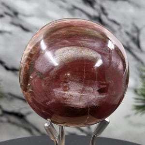 Puede incluir: Una piedra esférica pulida con una superficie lisa y brillante que presenta patrones de remolinos en tonos marrón rojizo profundo, crema y marrón oscuro. La esfera está sostenida por un soporte de metal plateado con tres finas varillas curvas.