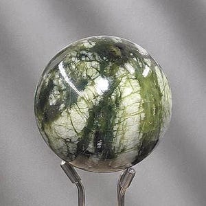 May include: A polished spherical stone featuring a marbled pattern of deep forest green and creamy white veins. The sphere is supported by a silver-toned metal stand with two prongs that cradle the base of the stone, providing a stable display.