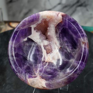 Könnte beinhalten: Eine polierte Amethystschale mit einem Geodenzentrum. Die Schale ist tiefviolett mit weißen und braunen Einschlüssen. Das Geodenzentrum hat eine kristalline Struktur. Die Schale ist rund und hat eine glatte, glänzende Oberfläche.