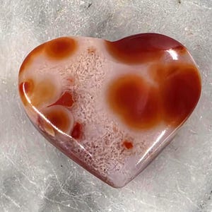 Puede incluir: Una piedra pulida en forma de coraz&oacute;n con un acabado brillante. La piedra presenta una mezcla de colores, incluyendo rojo intenso, naranja y blanco. El coraz&oacute;n mide aproximadamente 5 cm de ancho y tiene una forma suave y redondeada. El patr&oacute;n de la piedra es &uacute;nico.