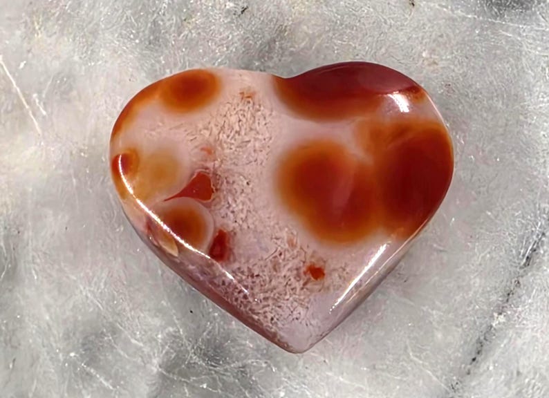 Puede incluir: Una piedra pulida en forma de coraz&oacute;n con un acabado brillante. La piedra presenta una mezcla de colores, incluyendo rojo intenso, naranja y blanco. El coraz&oacute;n mide aproximadamente 5 cm de ancho y tiene una forma suave y redondeada. El patr&oacute;n de la piedra es &uacute;nico.