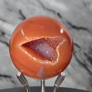 May include: A polished, orange-red agate sphere with a geode cavity. The cavity reveals a sparkling, crystalline interior with shades of purple and brown. The sphere is mounted on a silver-colored metal stand, showcasing the natural beauty of the gemstone.