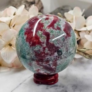 Könnte beinhalten: Eine polierte Edelstein-Kugel mit einem rot-grünen, gesprenkelten Muster, die auf einem kleinen, dunkelroten Ständer ruht. Die Kugel ist von weißen Blumen umgeben und bildet eine natürliche und dekorative Präsentation.
