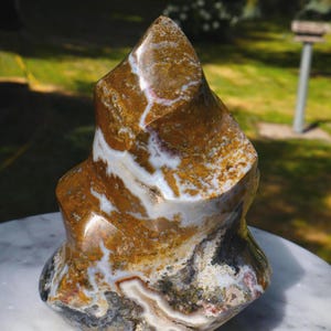 May include: A polished stone sculpture with an unusual, organic form. The stone displays a mix of brown, white, and grey hues, with complex patterns and textures. The sculpture is presented on a marble surface.