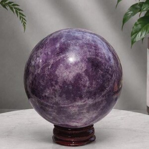 Peut inclure: Une boule de cristal en lépidolite violette polie et sphérique, présentant des inclusions minérales naturelles blanches et violet foncé, reposant sur un support de présentation circulaire en bois brun foncé à la surface lisse et réfléchissante.